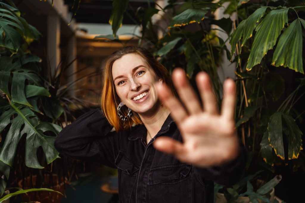 Anna Flemmer steht lächelnd zwischen grünen Pflanzen, eine Hand mit der Handfläche nah zur Kamera. 