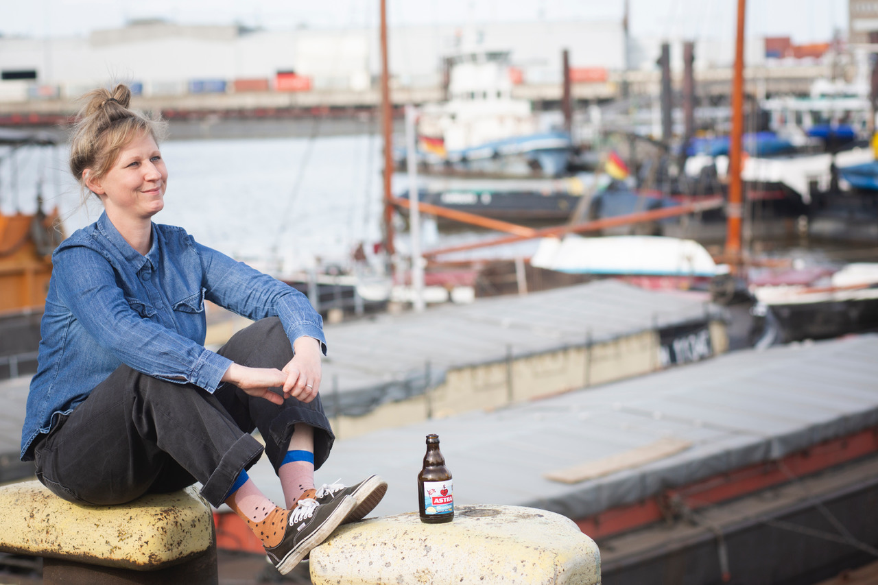 Martina sitzt entspannt auf einer Hafenmauer, im Hintergrund sieht man Schiffe